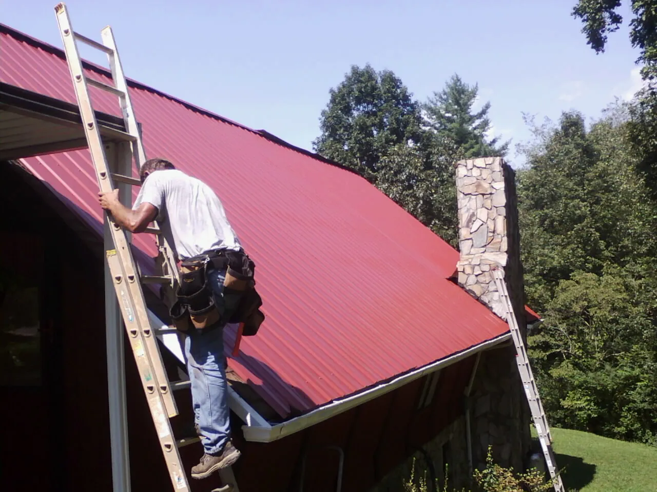 Roofer on a ladder beside a red metal roof and stone chimney — Soffit & Fascia Repair by Roofing Brothers in Nova